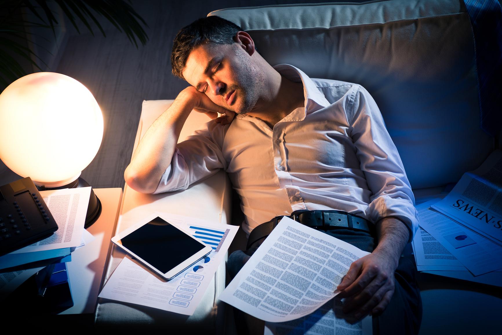 Tired businessman sleeping on sofa at home surrounded by paperwork.