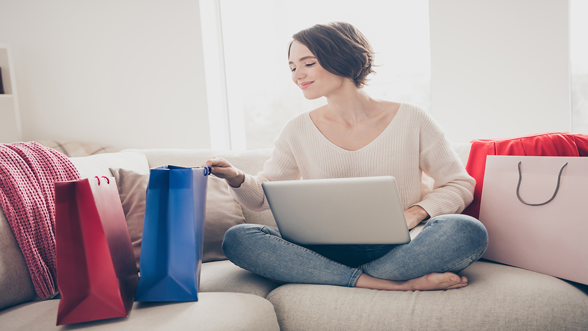 Full size photo of young woman look inside shopping bag hold laptop buy new clothes sit couch indoors in house