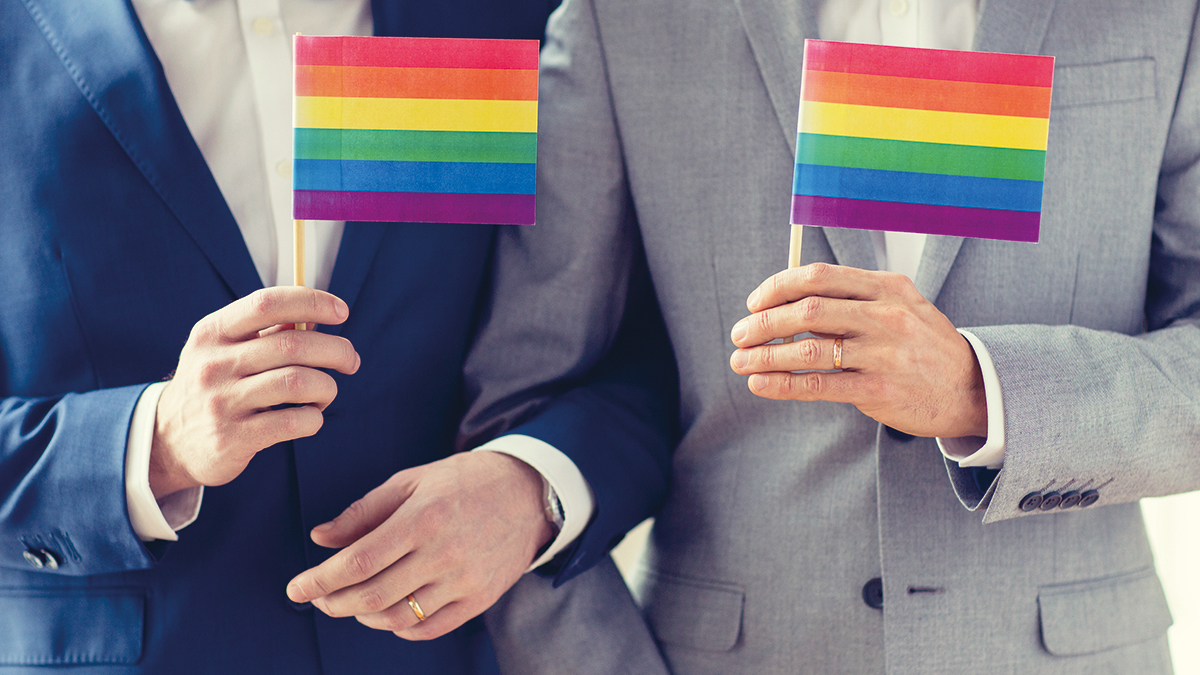 people, homosexuality, same-sex marriage and love concept – close up of happy male gay couple in suits and bow-ties with wedding rings holding rainbow flags