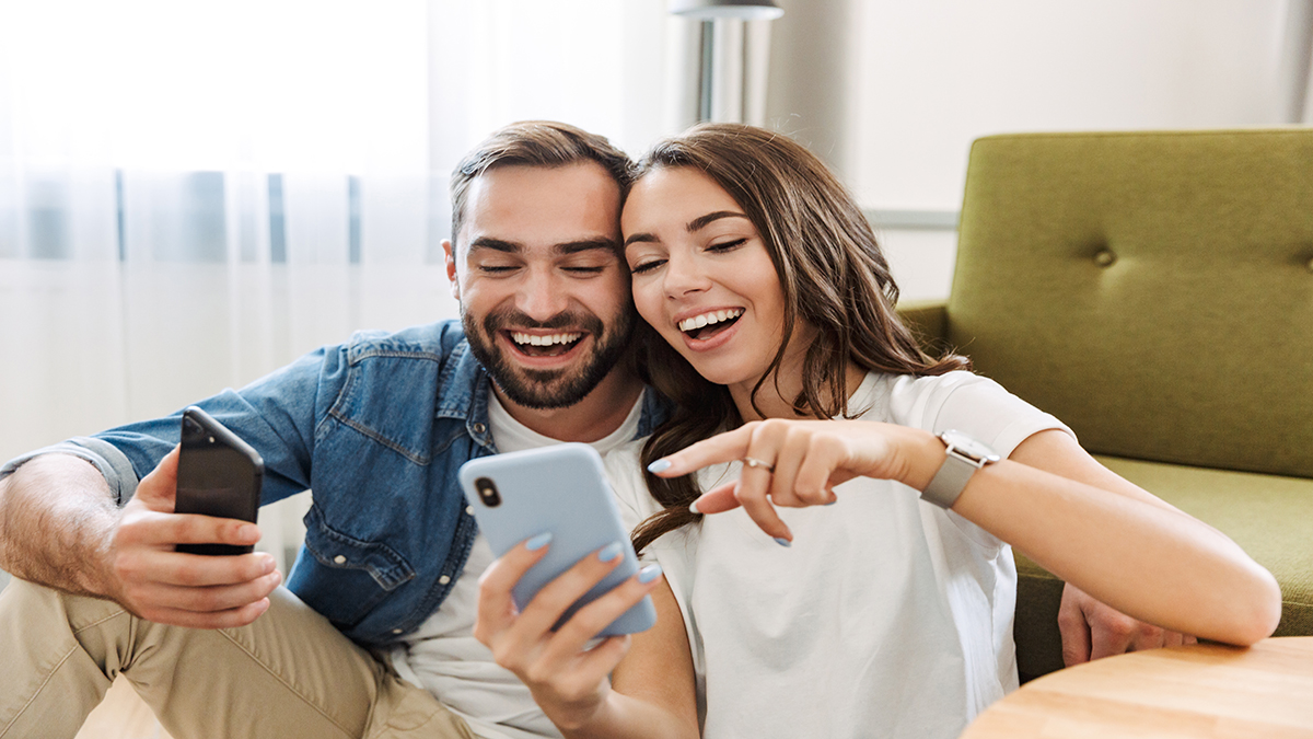 Beautiful young couple in love at home, sitting at the living room, pointing at mobile phone
