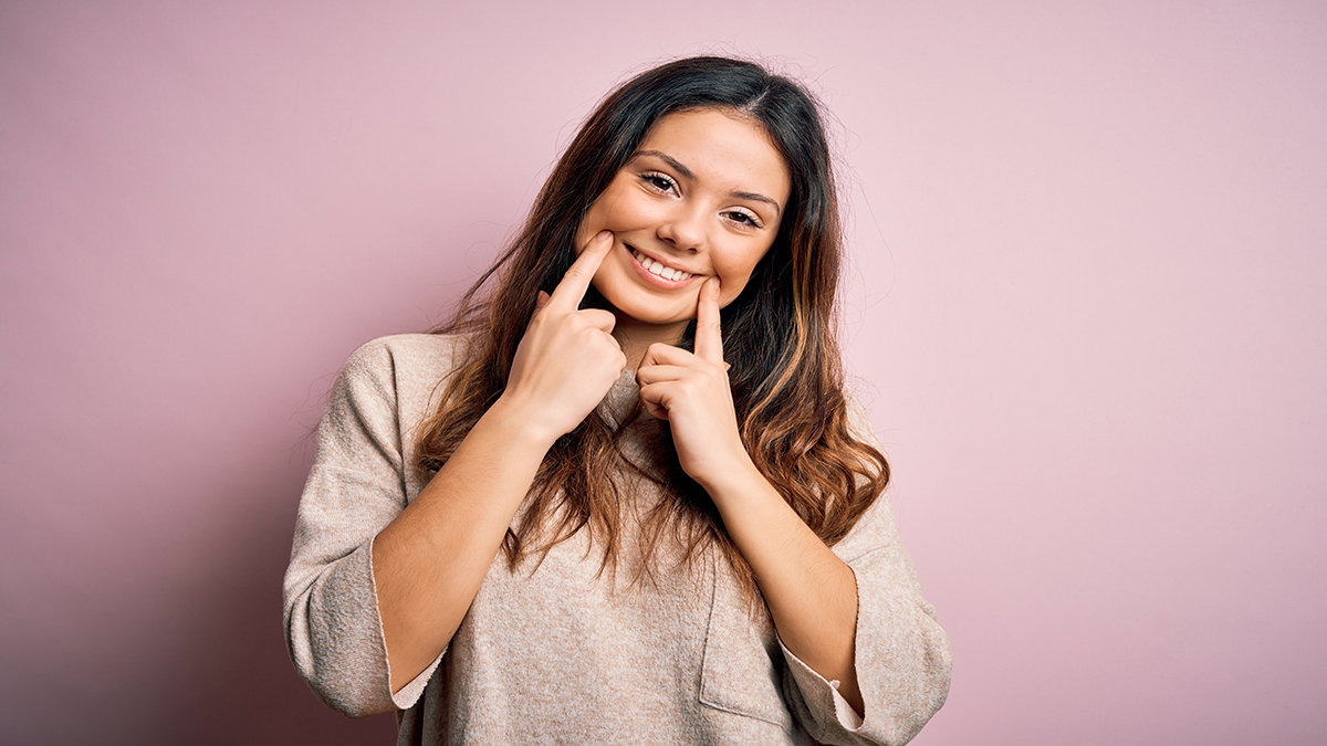 Young beautiful brunette woman wearing casual sweater standing over pink background Smiling with open mouth, fingers pointing and forcing cheerful smile